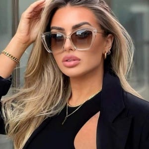 Pu&ograve; includere: Occhiali da sole con montatura trasparente e lenti sfumate marroni. La persona indossa un top e un blazer neri. Sono visibili anche una collana e un braccialetto dorati. Accessorio di moda.
