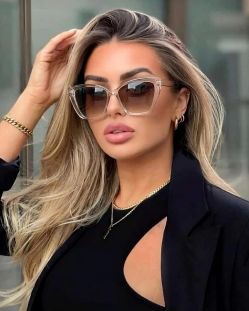 Pu&ograve; includere: Occhiali da sole con montatura trasparente e lenti sfumate marroni. La persona indossa un top e un blazer neri. Sono visibili anche una collana e un braccialetto dorati. Accessorio di moda.