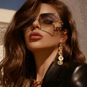 May include: A woman wearing oversized, rimless sunglasses with a gradient lens and gold accents. She is also wearing gold coin earrings and a gold chain necklace. The woman is wearing a black leather jacket.