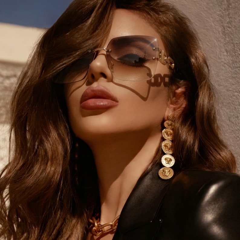 May include: A woman wearing oversized, rimless sunglasses with a gradient lens and gold accents. She is also wearing gold coin earrings and a gold chain necklace. The woman is wearing a black leather jacket.