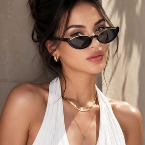 K&ouml;nnte beinhalten: Schwarze Cat-Eye-Sonnenbrille mit goldfarbenen Metallakzenten an den oberen R&auml;ndern. Die Fassung hat dunkel get&ouml;nte Gl&auml;ser und eine elegante, spitz zulaufende Form. Die Brille wird mit einem wei&szlig;en &auml;rmellosen Oberteil, goldenen Creolen und geschichteten Goldketten kombiniert.