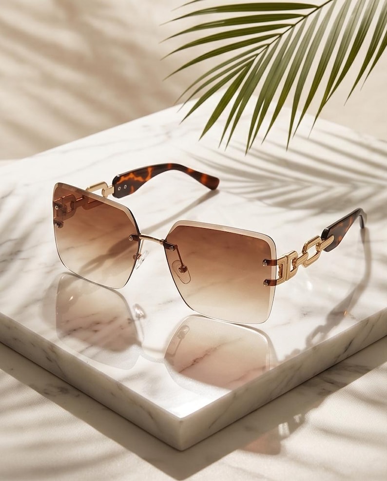 May include: Brown gradient sunglasses with a gold metal frame and chain detail on the temples. The sunglasses have a square shape and a tortoiseshell pattern on the arms. The lenses are a gradient brown color.