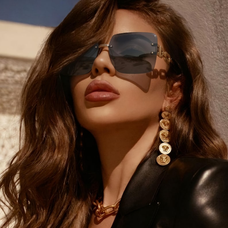 May include: Square-framed sunglasses with a gold metal frame and dark lenses. The sunglasses are worn with gold-toned earrings and a matching necklace. The image also shows a black leather jacket.