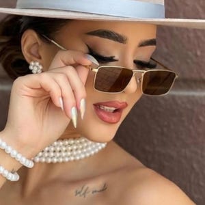 May include: Gold-framed sunglasses with brown lenses. The sunglasses are held up by a hand. The person is wearing a white hat, pearl jewelry, and has a tattoo that says "self made".