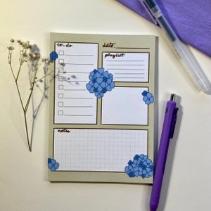 May include: A beige notepad with a floral design and sections for to-do lists, dates, playlists, and notes. Blue hydrangea illustrations accent the corners and center. A purple pen and a clear pen are placed on the side.