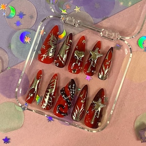 May include: A set of red and silver press-on nails in a clear plastic case. The nails feature various designs, including silver accents, star and heart embellishments, and a butterfly design. The nails are long and pointed.