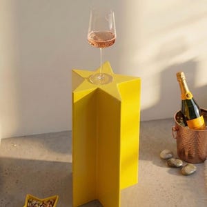 May include: A tall, bright yellow star-shaped pedestal stand with a flat top surface. A clear glass filled with a pink beverage rests on the pedestal. A small yellow star-shaped bowl filled with mixed nuts sits nearby on the surface.