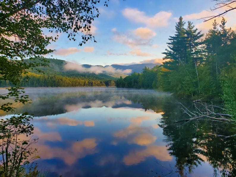 May include: A serene landscape featuring a calm lake reflecting the sky and surrounding trees. The sky is a mix of blue and peach hues, with fluffy clouds mirrored in the water. Lush green trees line the lake's edge, creating a peaceful natural scene.