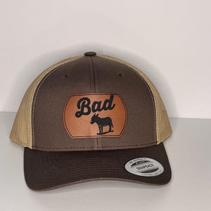 May include: Brown and tan trucker hat with a leather patch. The patch features the word "Bad" above a silhouette of a donkey. The hat has a curved brim and a snapback closure.