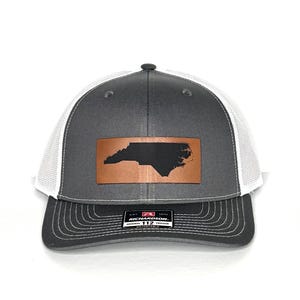 May include: Gray and white trucker hat with a leather patch featuring a black outline of North Carolina. The hat has a curved brim and an adjustable strap. The Richardson brand label is visible.