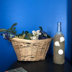 May include: A wicker basket filled with white and purple flowers sits on a black surface next to a clear glass bottle decorated with white seashells. The bottle is filled with sand and seashells. The text "Every pretty pirate needs a beautiful mermaid" is on a coaster in the foreground.