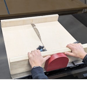 Puede incluir: Primer plano de una sierra de mesa con una valla de madera y un empujador rojo. La hoja de sierra es visible y la valla está configurada para cortar una pieza de madera.