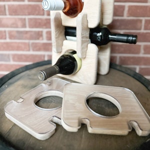 May include: A wooden wine rack holding three wine bottles, with two matching coasters in front. The rack is made of light-colored wood and has a castle-like design. The bottles are filled with red and white wine.