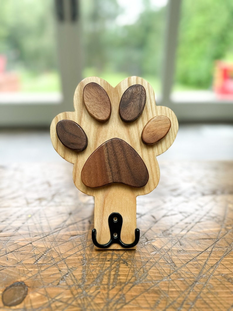 May include: Wooden paw print shaped coat hook with a black metal double hook. The paw print is made of light wood with darker wood accents.