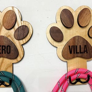 May include: Two wooden dog paw-shaped leash holders with the names "HERO" and "VILLA" engraved on them. Each holder has a leash attached to it, one teal and one pink.