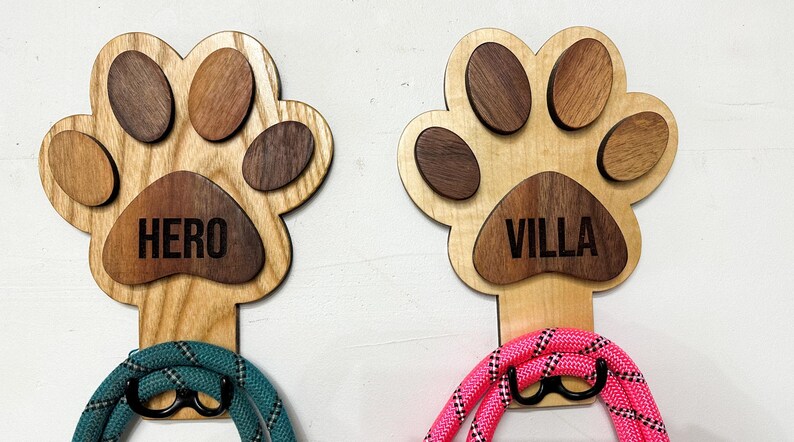 May include: Two wooden dog paw-shaped leash holders with the names "HERO" and "VILLA" engraved on them. Each holder has a leash attached to it, one teal and one pink.