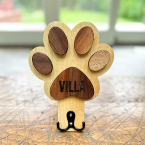 May include: Wooden dog paw print coat hook with the name "VILLA" engraved on the bottom. The paw print is made of different shades of wood.