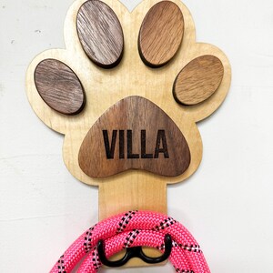 May include: Wooden paw print dog leash hook with the name "VILLA" engraved on it. The hook is painted in a light wood color and has a pink leash hanging from it.