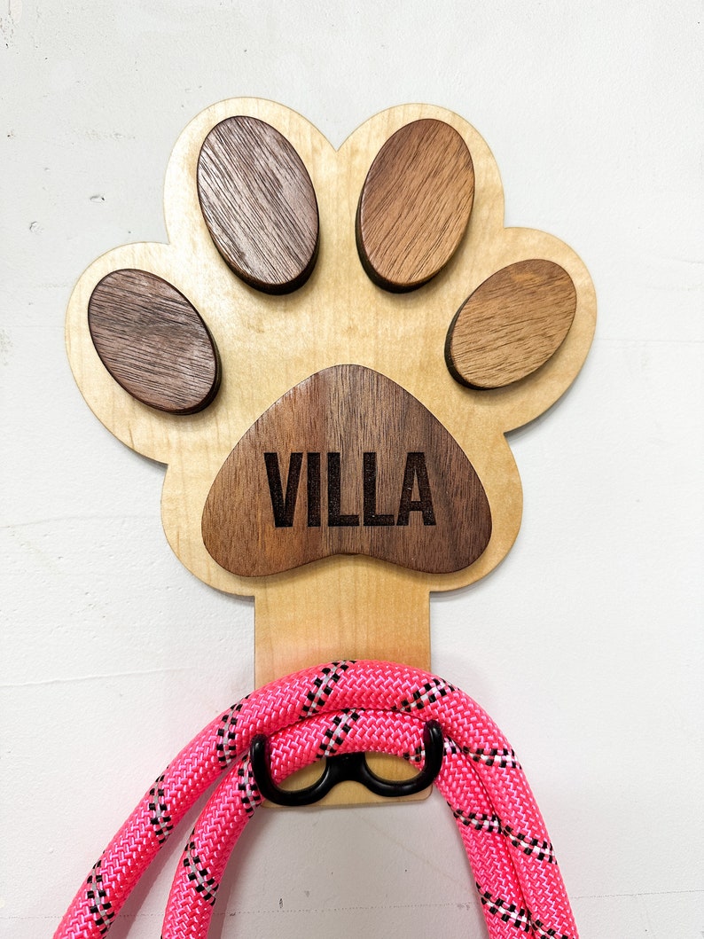 May include: Wooden paw print dog leash hook with the name "VILLA" engraved on it. The hook is painted in a light wood color and has a pink leash hanging from it.