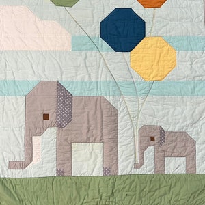 Puede incluir: Una manta de bebé acolchada con un elefante gris y un bebé elefante sosteniendo globos de colores. Los globos son verdes, naranjas, azules y amarillos. El fondo es azul claro con nubes blancas.