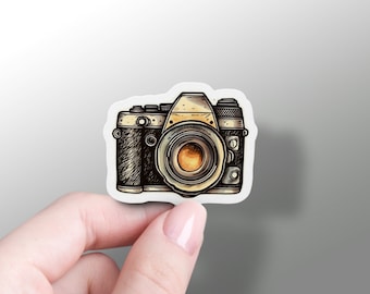 May include: A vintage-style camera sticker with a black and brown body, a gold lens, and a white background.