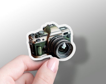 May include: A vintage camera sticker with a green and beige design. The camera has a large lens with the text "C.I.ENON 1:1.8" printed on it.