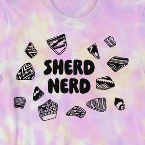 Puede incluir: Una camiseta tie-dye rosa, amarilla y blanca con un gráfico en blanco y negro de fragmentos de cerámica y el texto "SHERD NERD".