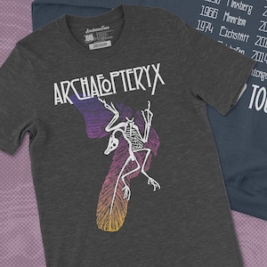 Peut inclure: Un t-shirt gris foncé avec un graphique blanc d'un squelette d'Archaeopteryx, un oiseau préhistorique, avec le texte "ARCHAEOPTERYX" au-dessus. Un sweat-shirt bleu foncé avec le texte "LIFT-OFF" et une liste de villes et d'années est visible en arrière-plan.