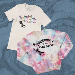 Op de afbeelding: Twee T-shirts met een tie-dye ontwerp. Het witte T-shirt heeft de tekst "Archeologie is mijn PASSIE" in regenboogkleuren. Het tie-dye T-shirt heeft dezelfde tekst in zwart. Beide T-shirts hebben een schedelgrafiek.