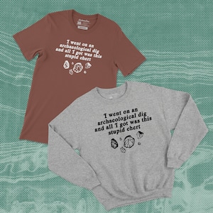 Może przedstawiać: Dwie koszulki, brązowy t-shirt z krótkim rękawem i szara bluza z długim rękawem, obie z napisem "I went on an archaeological dig and all I got was this stupid chert" oraz grafikami krzemienia.
