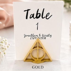 May include: Five table number cards with the text Table 1, Jonathan & Kirsty, and 23/07/2011, each held by a triangular metal stand featuring a circle and vertical line design. The stands are available in gold, silver, black, white, and copper finishes.