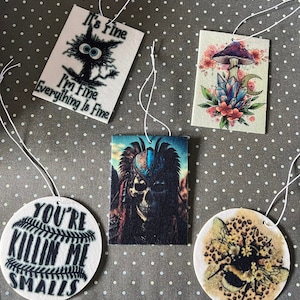 May include: Five different car air fresheners with various designs. One features a black cat with the text "It's fine I'm fine Everything is fine". Another features a mushroom with crystals and a crescent moon. A third features a skull with a headdress. A fourth features a baseball with the text "You're killin' me Smalls". The last one features a bee with flowers and a leopard print background.