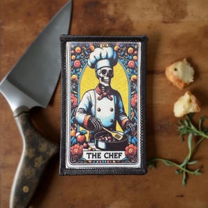 May include: A rectangular patch featuring a skeleton chef in a white uniform and chef's hat, cooking in a pan. The design is framed with floral and fruit motifs, with the text "THE CHEF" at the bottom. The patch is on a wooden surface with a knife and food.