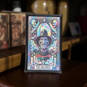 May include: A rectangular patch with a black border featuring a colorful illustration of a character wearing a hat and striped sweater. The image includes skulls, candles, and the text "THE DREAMER".