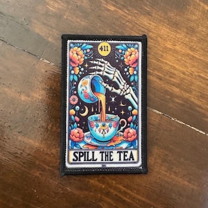 May include: Rectangular patch with a black border featuring a colorful illustration. A skeleton hand pours liquid from a decorated cup into a teacup. The text "SPILL THE TEA" is at the bottom, with the number "411" at the top.