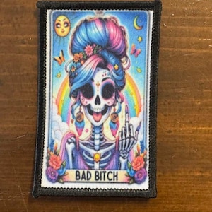 May include: A rectangular patch featuring a colorful illustration of a sugar skull with blue, pink, and purple hair, a rainbow, and the text "BAD BITCH". The patch has a black border and is likely for clothing or accessories.