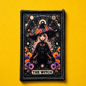 May include: A rectangular embroidered patch featuring a central illustration of a figure with long light hair wearing a wide-brimmed hat adorned with colorful flowers. The design includes a black dog, celestial motifs, and the text THE WITCH.