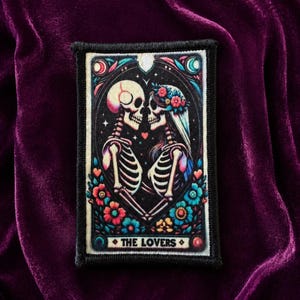May include: A rectangular embroidered patch featuring a tarot card design titled THE LOVERS. The artwork depicts two skeletons facing each other, surrounded by colorful flowers, a heart, and celestial motifs in shades of pink, blue, and white.