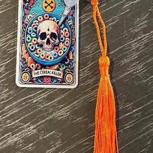 May include: A novelty keychain featuring a tarot card design with a skull in a bowl of cereal, the text "THE CEREAL KILLER", and an orange tassel. The keychain is attached to a silver keyring.