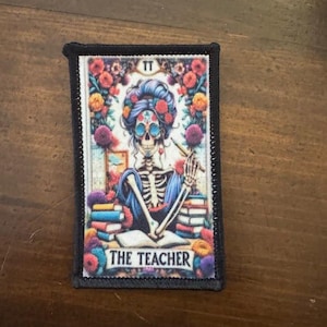 May include: Rectangular patch with a Day of the Dead-style illustration of a skeleton teacher. The image features a skeleton with a floral headdress, surrounded by flowers and books. The text "THE TEACHER" is at the bottom.