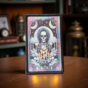May include: A rectangular patch with a black border features a colorful illustration of a skeleton with blue hair, wearing a striped suit. The image includes moons, clouds, candles, and the text "THE TRICKSTER". The patch is displayed on a wooden surface.