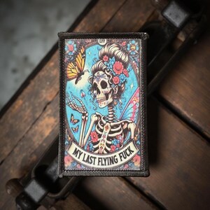 May include: Rectangular patch featuring a skeleton fairy with a butterfly and floral accents. The patch has the text "MY LAST FLYING FUCK" on a banner. The design is set against a blue background with a black border.