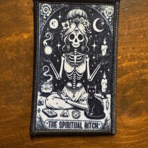 May include: A black and white patch featuring a skeleton figure in a meditative pose, adorned with floral elements and celestial symbols. A black cat sits beside the figure. The text "THE SPIRITUAL BITCH" is at the bottom.