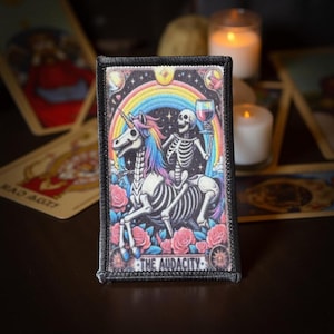 May include: Rectangular patch featuring a skeleton riding a skeleton unicorn under a rainbow. The unicorn has a colorful mane and tail. The skeleton holds a glass of wine. Pink roses surround the image, with the text "THE AUDACITY" at the bottom.