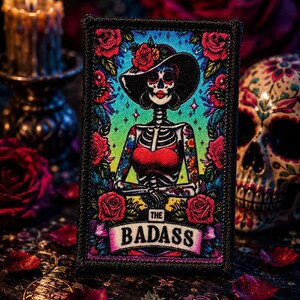 May include: A rectangular embroidered patch featuring a skeleton figure wearing a wide-brimmed hat and a red top, surrounded by vibrant red roses and a colorful gradient background. The text THE BADASS is printed on a banner at the bottom.