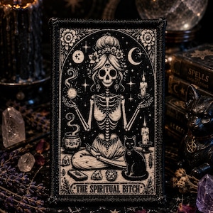 May include: A rectangular embroidered patch featuring a black and white illustration of a skeleton sitting in a lotus position, surrounded by candles, a cat, and celestial symbols, with the text THE SPIRITUAL BITCH printed at the bottom.
