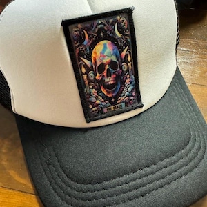 Peut inclure: Casquette de camionneur noire et blanche avec un écusson représentant un crâne coloré. L'écusson porte l'inscription "THE DEATH" et se détache sur un fond de bougies et d'éléments célestes. La casquette a une visière noire et un panneau avant blanc.