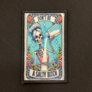 May include: Rectangular patch with a black border featuring a skeleton pouring salt from a shaker. The text "DON'T BE" is above the skeleton, and "A SALTY BITCH" is below. The background is turquoise with floral accents.