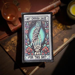 May include: A rectangular patch with the text "NOT ENOUGH SAGE FOR THIS SHIT" features a skeleton hand holding a bundle of sage. The design includes red roses, purple leaves, and a teal background, all framed by a black border.