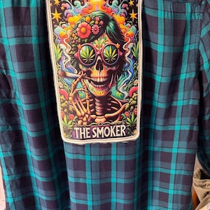 May include: A blue and green plaid flannel shirt with a patch featuring a colorful skull wearing sunglasses with marijuana leaves. The skull is smoking a cigarette, with the text "THE SMOKER" below.